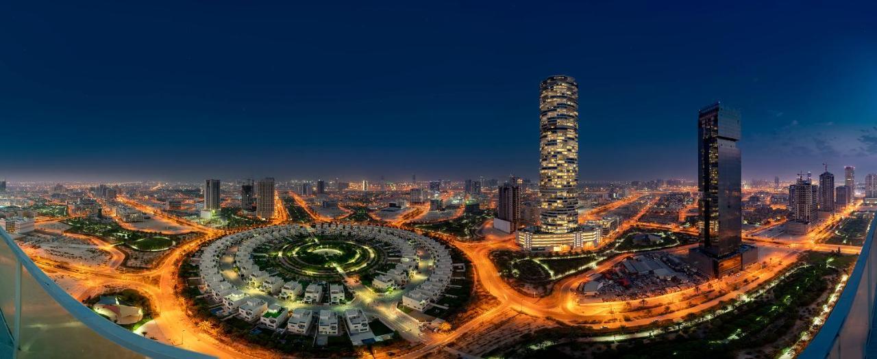 Five Jumeirah Village ดูไบ ภายนอก รูปภาพ The photo showcases a cityscape illuminated at night. In the foreground, there is a circular arrangement of buildings or houses, creating a visually striking pattern. To the right, a tall, modern skyscraper stands out, featuring a sleek design with n