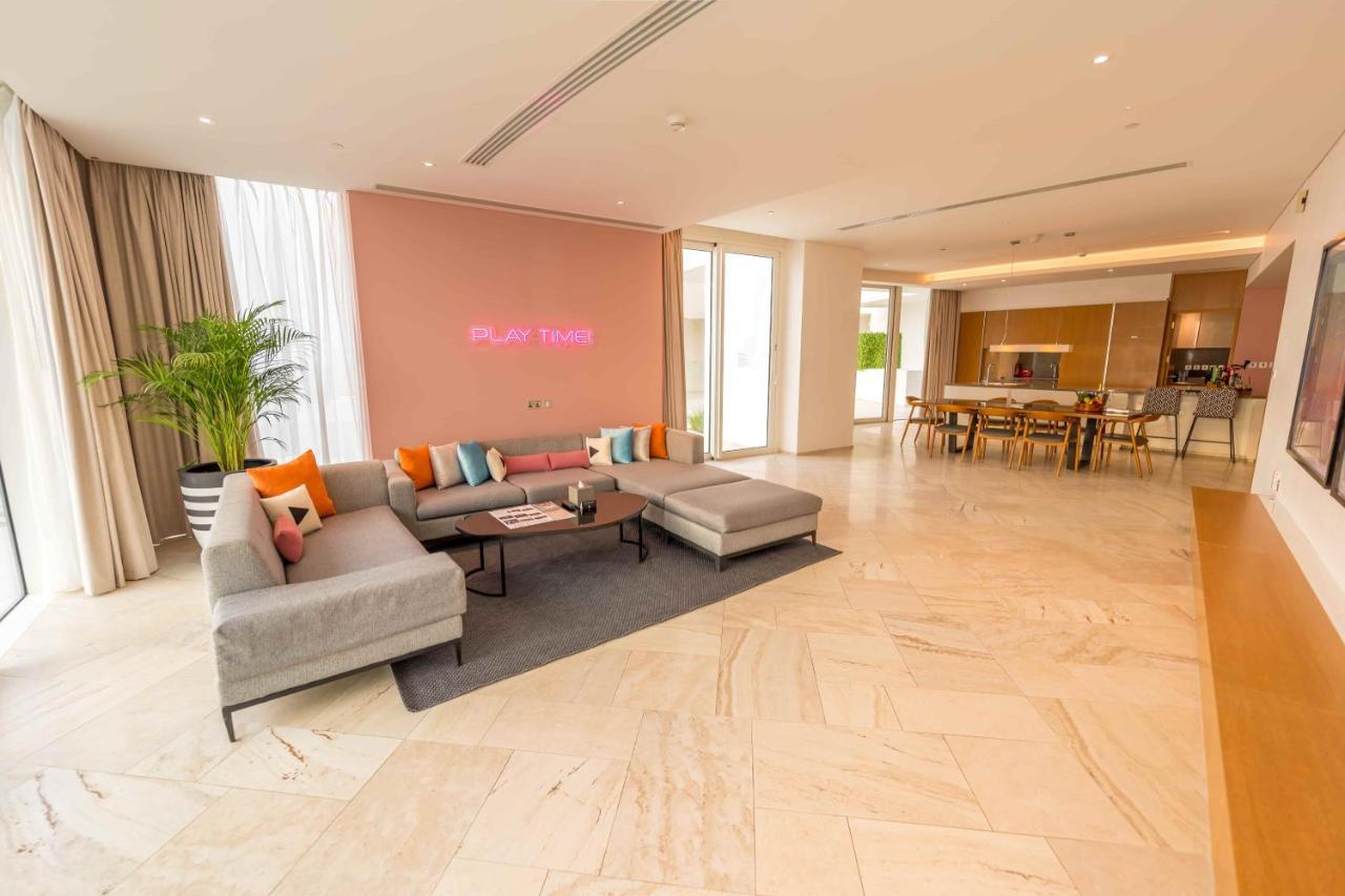 Five Jumeirah Village ดูไบ ภายนอก รูปภาพ The photo shows a spacious and modern living area. There is a large, light-colored sectional sofa with colorful cushions, positioned on a dark rug. The wall behind the sofa is painted in a soft pink shade, and there is a subtle decorative element or 