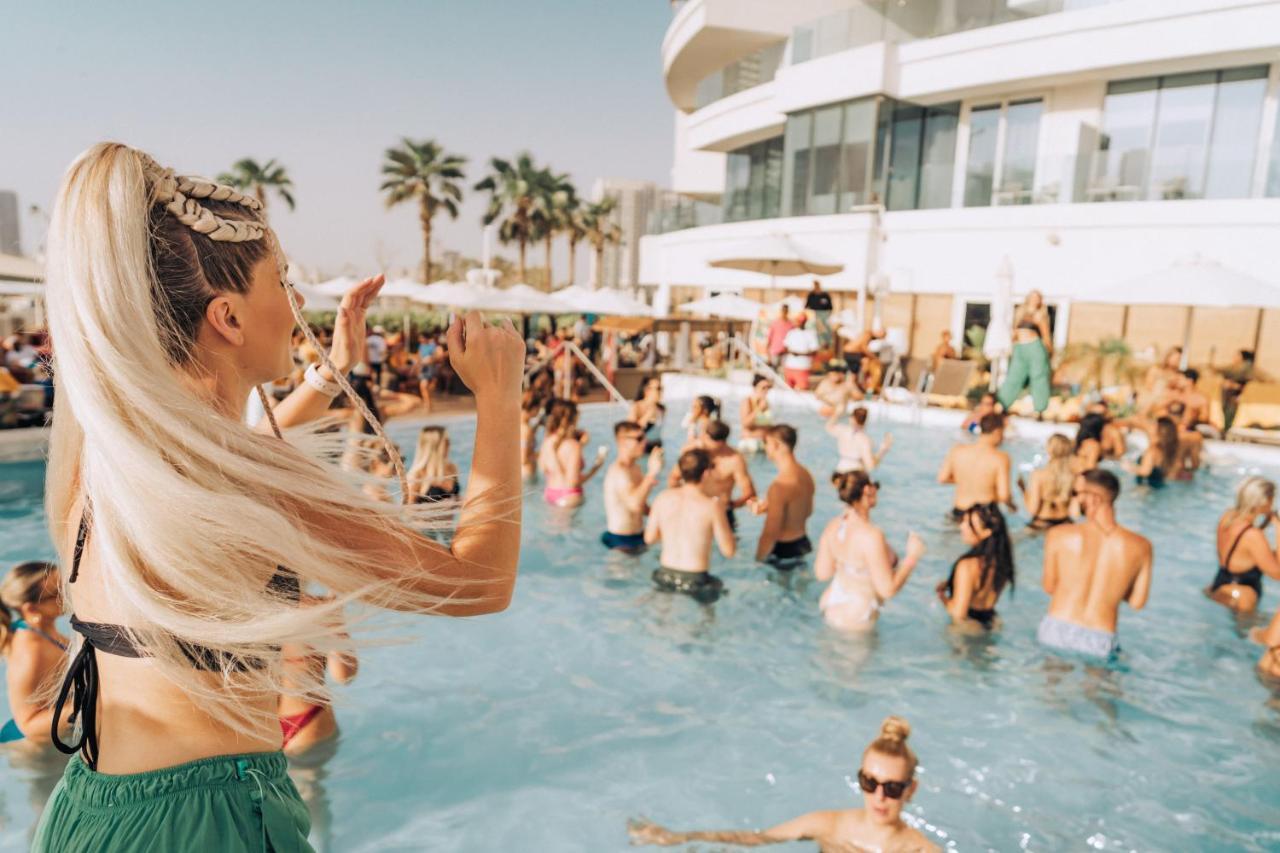 Five Jumeirah Village ดูไบ ภายนอก รูปภาพ The photo features a lively pool party scene. In the foreground, a woman with long, blonde hair is seen raising her arm as if enjoying the moment. The pool is filled with many people, some swimming and others socializing. The atmosphere appears festi