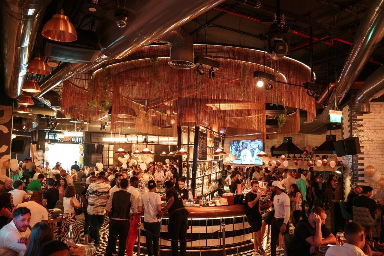Five Jumeirah Village ดูไบ ภายนอก รูปภาพ The photo depicts a lively bar or restaurant scene filled with people socializing. The interior design features a modern aesthetic with a mix of industrial elements, such as exposed pipes and metallic accents. A large, circular bar is the focal point
