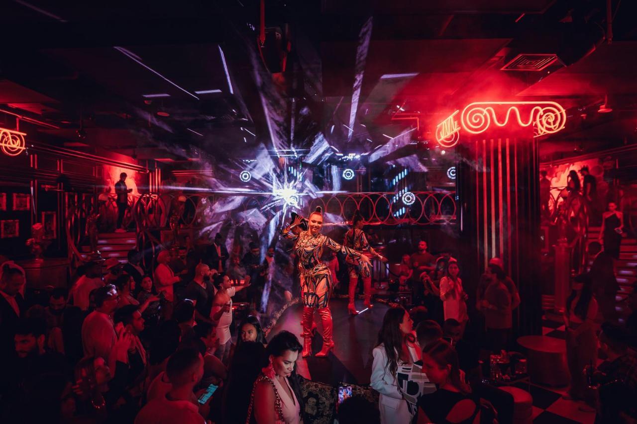 Five Jumeirah Village ดูไบ ภายนอก รูปภาพ The photo depicts a vibrant nightlife scene, likely from a nightclub or performance venue. The setting is dimly lit with striking red lights and dynamic visual effects, creating an electrifying atmosphere. In the center, a performer dressed in a flas