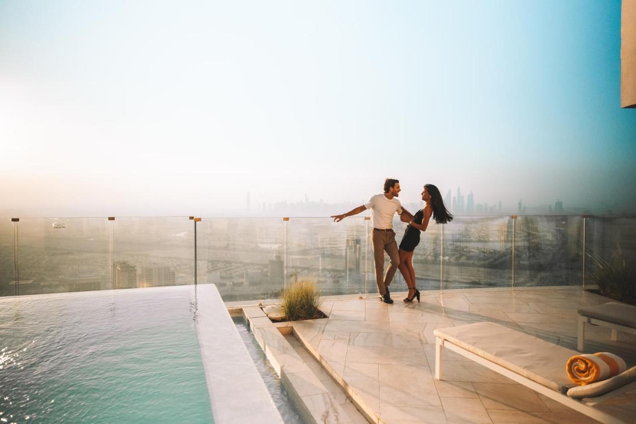 Five Jumeirah Village ดูไบ ภายนอก รูปภาพ The image shows a couple dancing together on a terrace or balcony with a view of a city skyline in the background. They appear to be enjoying a romantic moment, surrounded by a luxurious setting that includes a swimming pool. The atmosphere is bright
