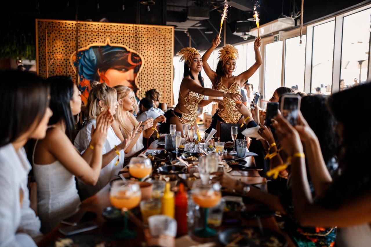 Five Jumeirah Village ดูไบ ภายนอก รูปภาพ The photo depicts a lively restaurant scene where two performers are entertaining a group of seated diners. The performers, dressed in colorful and elaborate outfits, are holding sparklers, adding a festive atmosphere to the occasion. The diners, mos