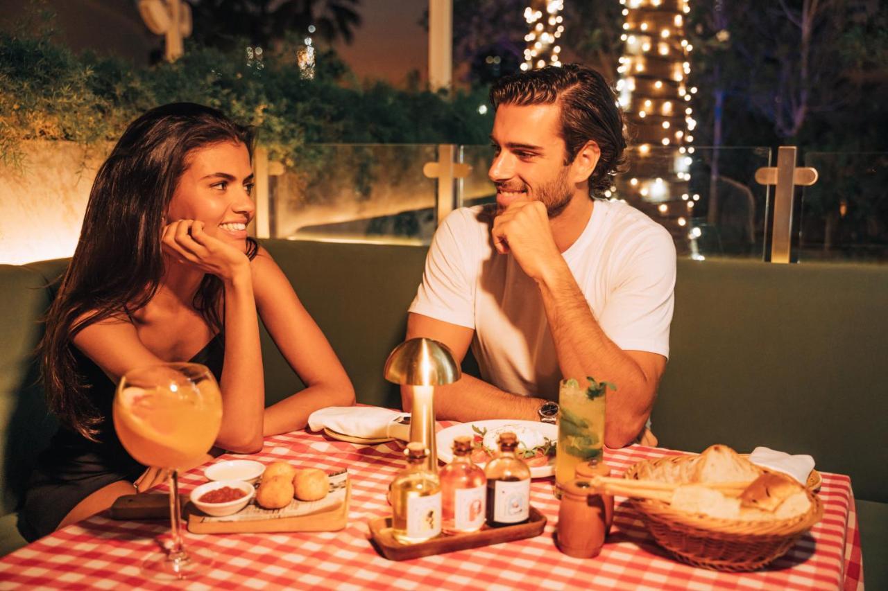 Five Jumeirah Village ดูไบ ภายนอก รูปภาพ The photo shows a romantic dining scene featuring a couple at a table. The setting is illuminated with soft lights, creating a warm atmosphere. The table is adorned with a red and white checkered tablecloth, and various dishes are presented, includin
