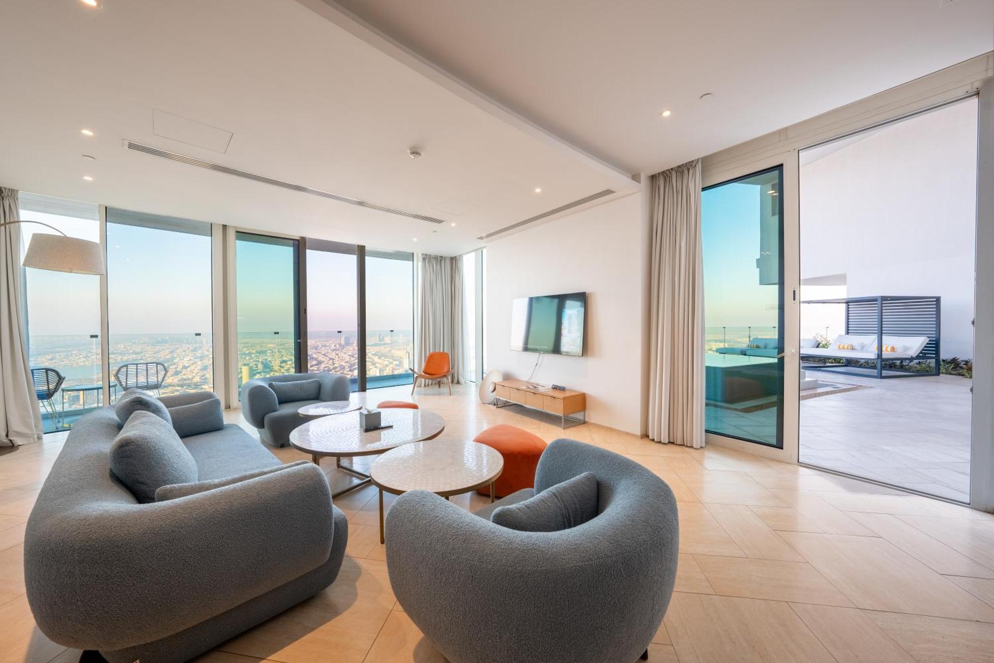 Five Jumeirah Village ดูไบ ภายนอก รูปภาพ The photo shows a modern and stylish living room area with large windows that provide a scenic view. There are several rounded, gray sofas arranged around a couple of round coffee tables. A sleek television is mounted on the wall across from the seat