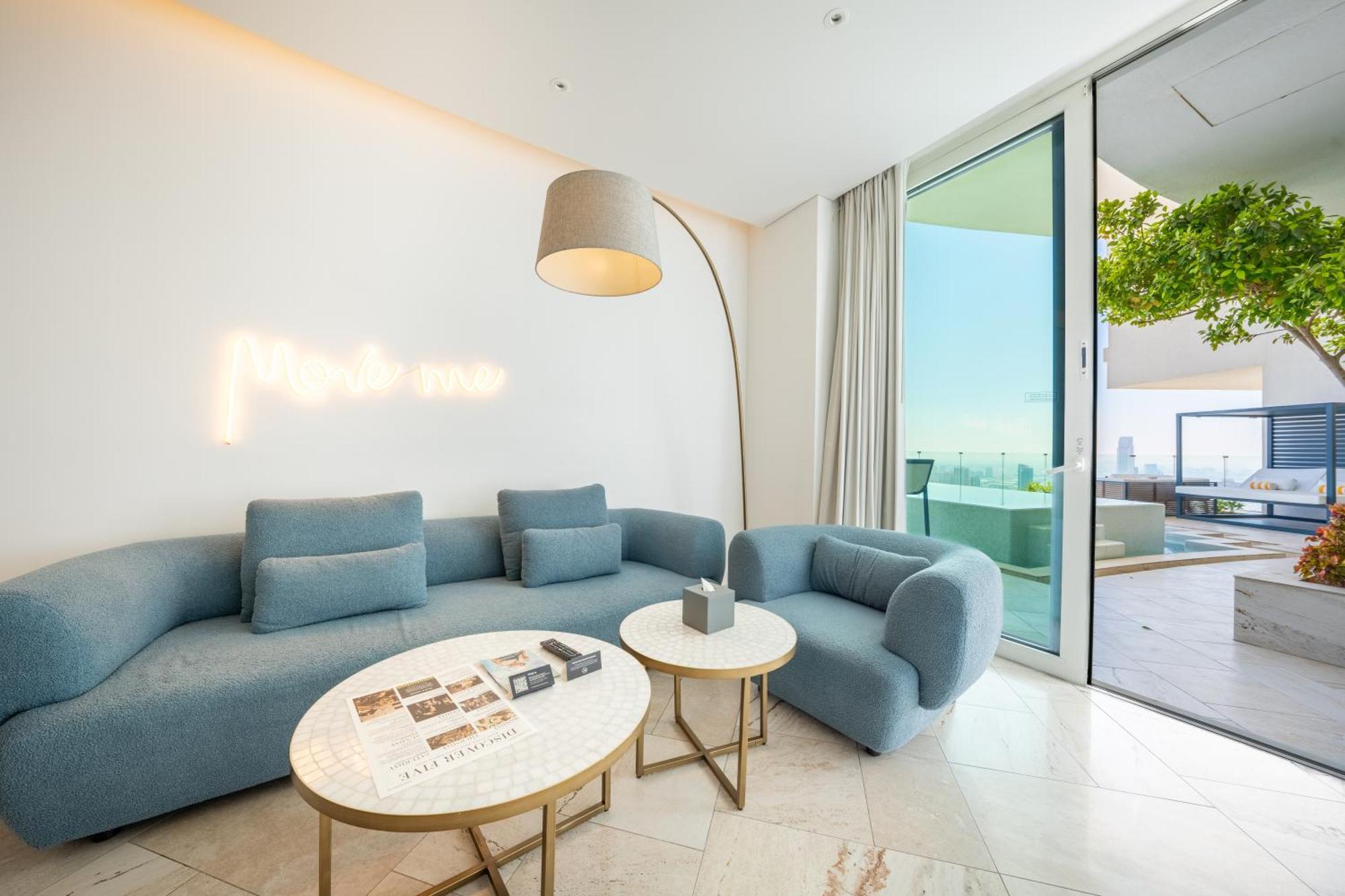 Five Jumeirah Village ดูไบ ภายนอก รูปภาพ The photo shows a modern and stylish living space. It features a light blue sectional sofa and a matching armchair, creating a cozy seating area. Two round marble coffee tables are placed in front, one of which has some magazines or books on it. A ta