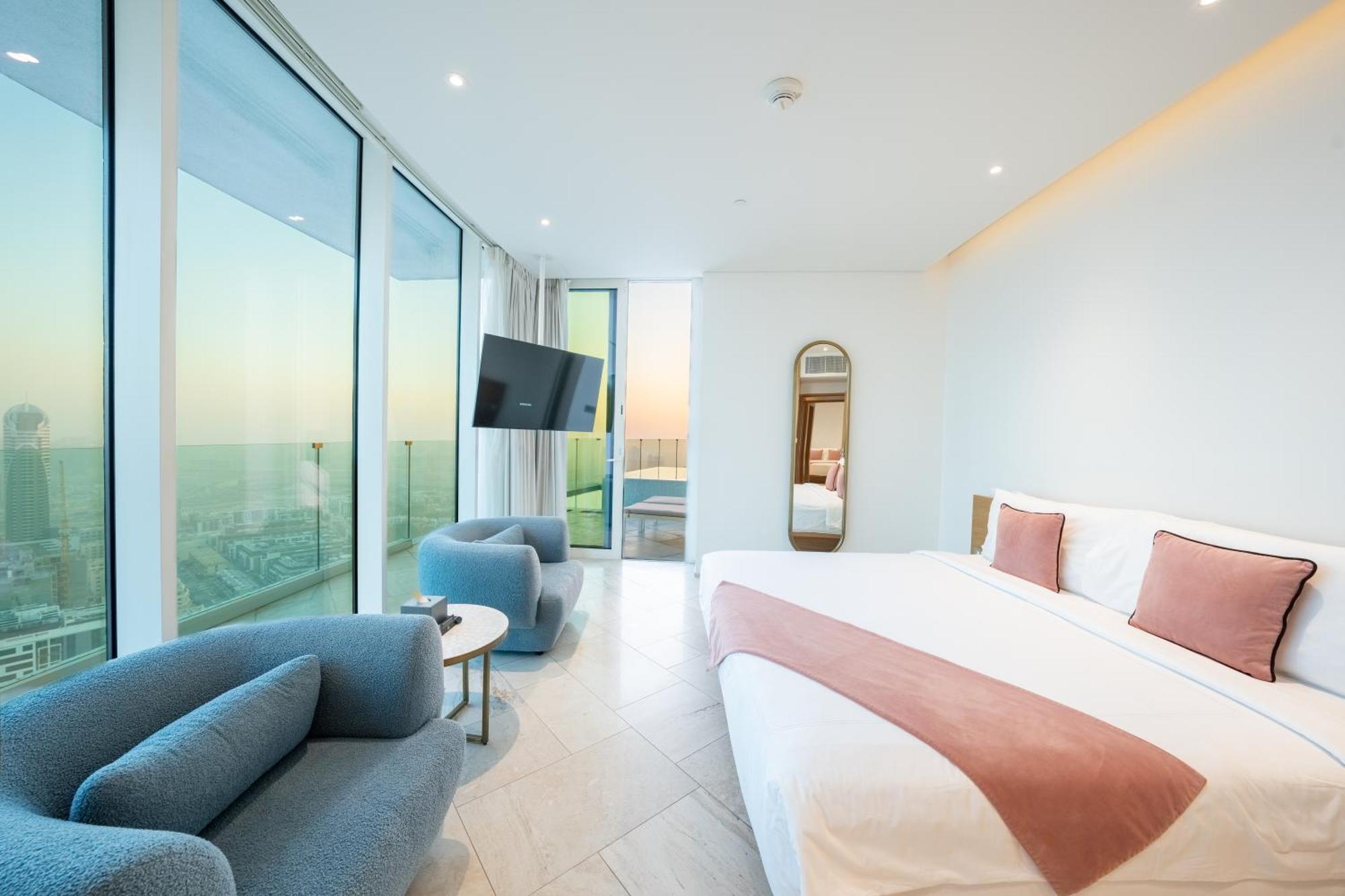 Five Jumeirah Village ดูไบ ภายนอก รูปภาพ The photo depicts a modern hotel room featuring large windows that provide a view of the outside. The room includes a spacious bed with a pink accent blanket, a cozy blue sofa, a small round table, and a wall-mounted television. There is also a mirro