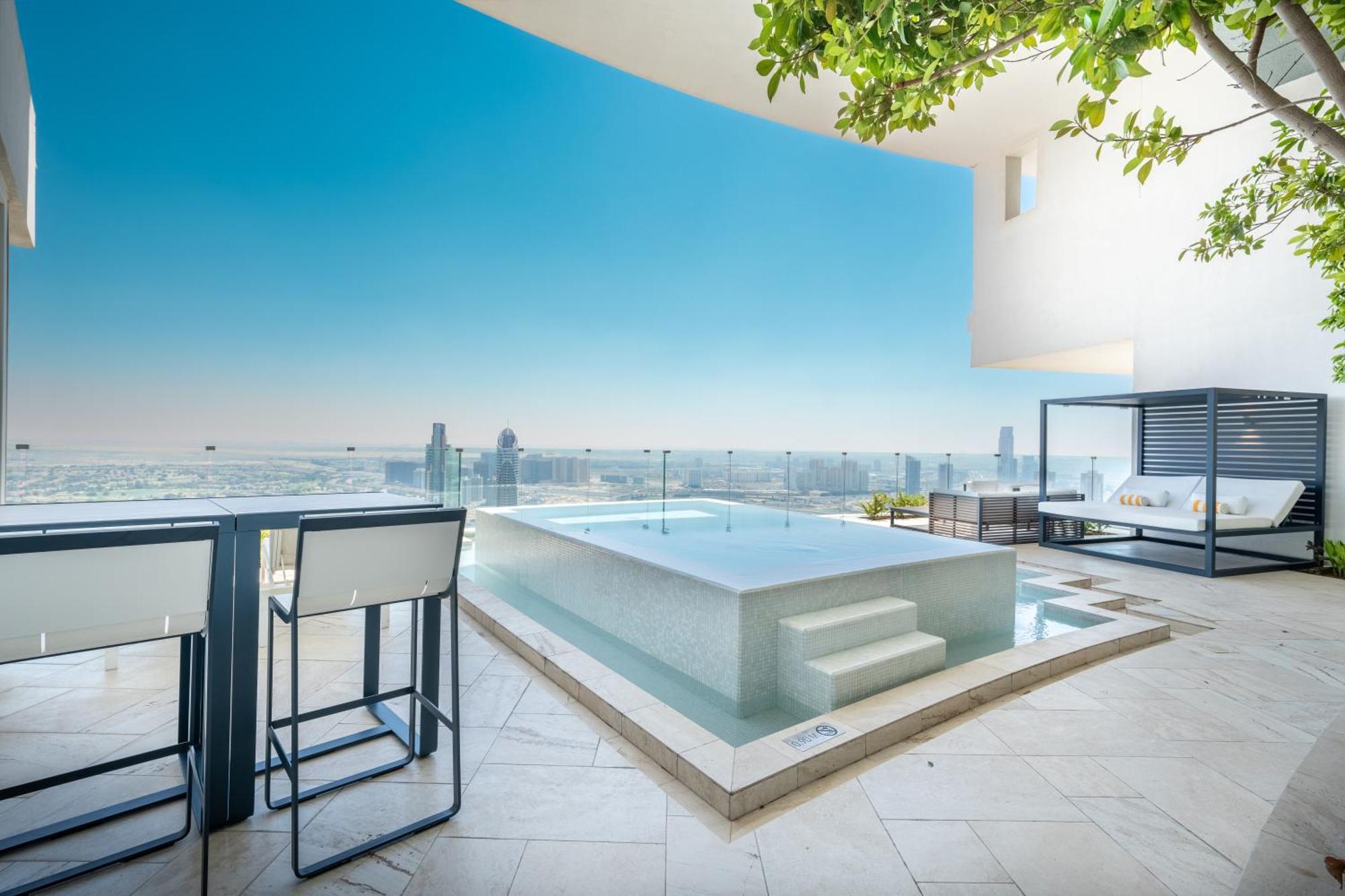 Five Jumeirah Village ดูไบ ภายนอก รูปภาพ The photo shows a modern outdoor space featuring a sleek infinity pool. The pool appears to be situated on a terrace with a stunning view of a city skyline or landscape in the background. There are elegant lounge chairs and a shaded cabana area, crea
