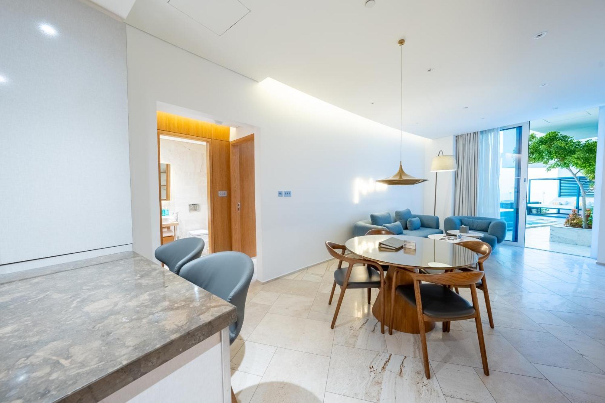 Five Jumeirah Village ดูไบ ภายนอก รูปภาพ The photo shows a modern living space that combines a dining area and a lounging area. On the left, there is a kitchen counter with a marble surface and several gray chairs around it. In the center, there is a round dining table made of wood with sev
