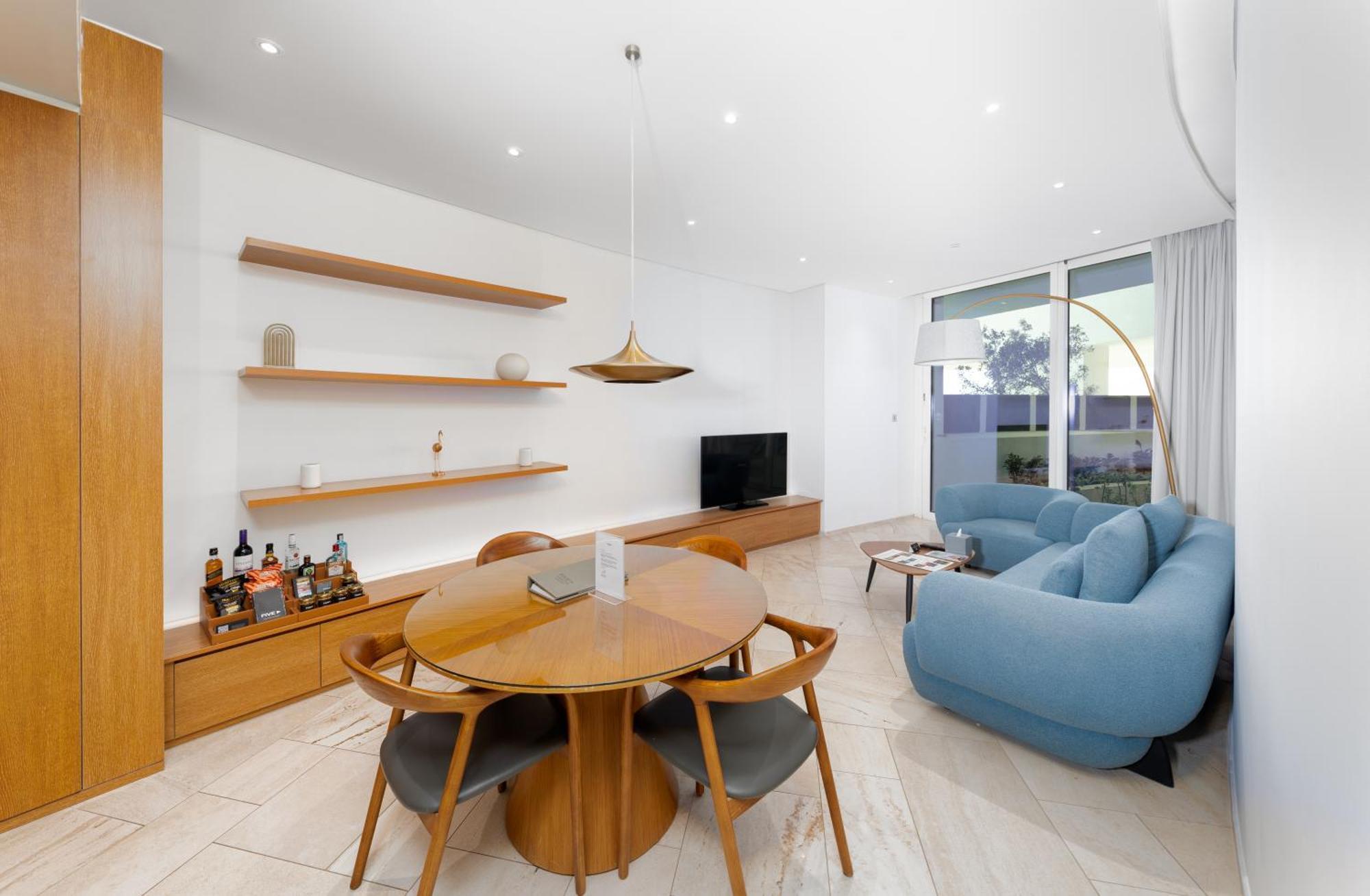 Five Jumeirah Village ดูไบ ภายนอก รูปภาพ The image shows a modern living space that features a cozy and stylish design. On one side, there is a round wooden dining table surrounded by four chairs. The table is set with a small item, possibly a decorative piece or a book. 

A pendant light h
