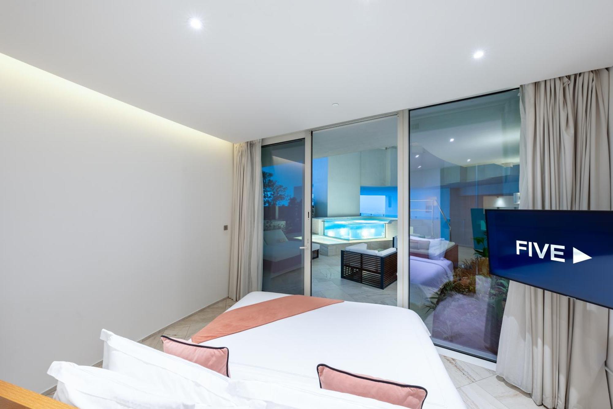 Five Jumeirah Village ดูไบ ภายนอก รูปภาพ The photo shows a modern, stylish bedroom featuring a large bed with light-colored bedding and decorative pillows. There are large glass windows that open up to a view of an outdoor area, likely overlooking a pool or patio. The room has neutral-color