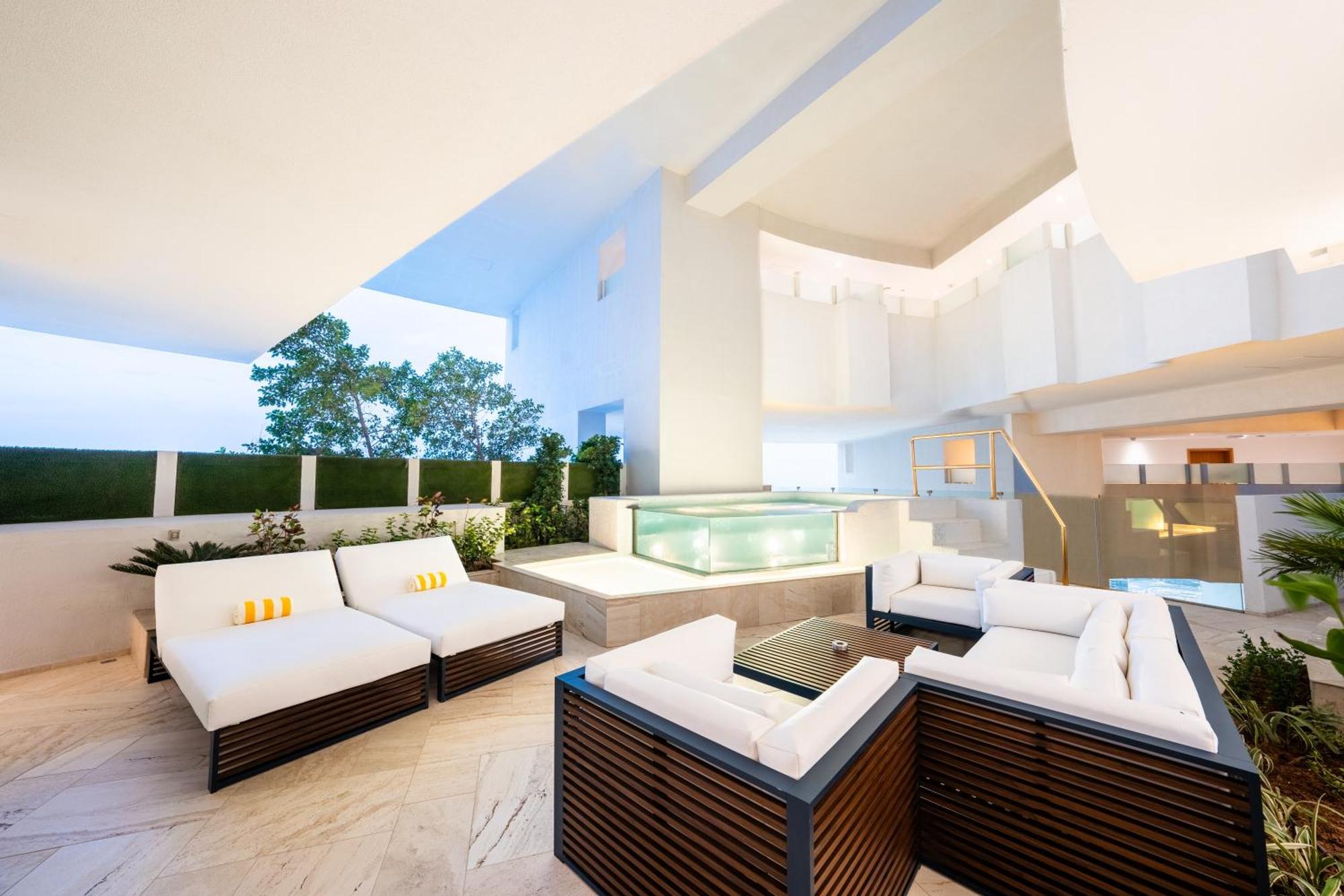 Five Jumeirah Village ดูไบ ภายนอก รูปภาพ The photo depicts a modern outdoor lounge area featuring comfortable seating arrangements. There are several white sofas with dark wooden frames, arranged around a glass coffee table. In the background, there is a sleek water feature or pool, surroun