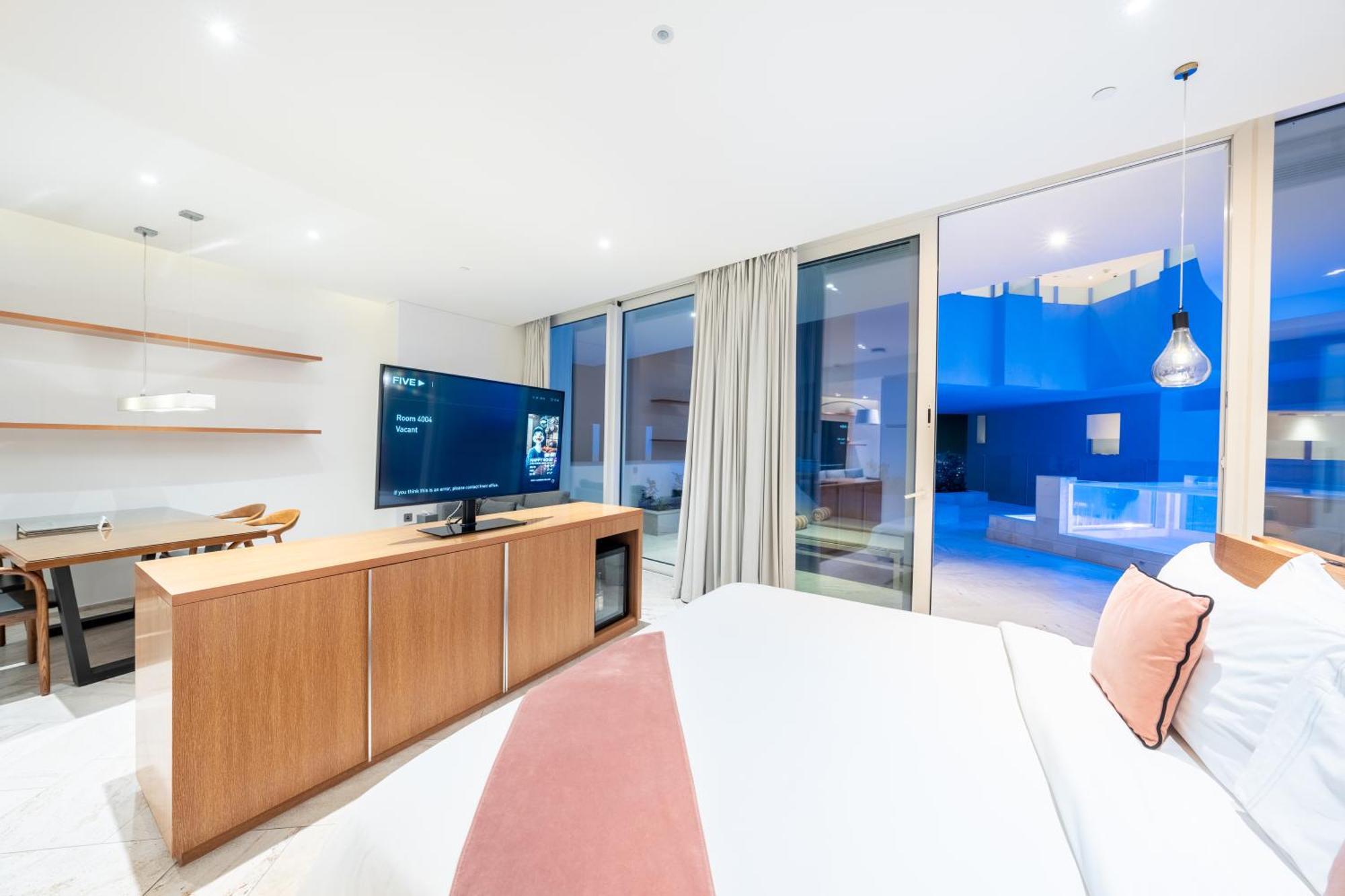 Five Jumeirah Village ดูไบ ภายนอก รูปภาพ The photo depicts a modern hotel room with a clean and minimalist design. A large bed is shown, topped with white linens and decorative pillows. Opposite the bed, there is a wooden TV stand with a flat-screen television. The room features large windo
