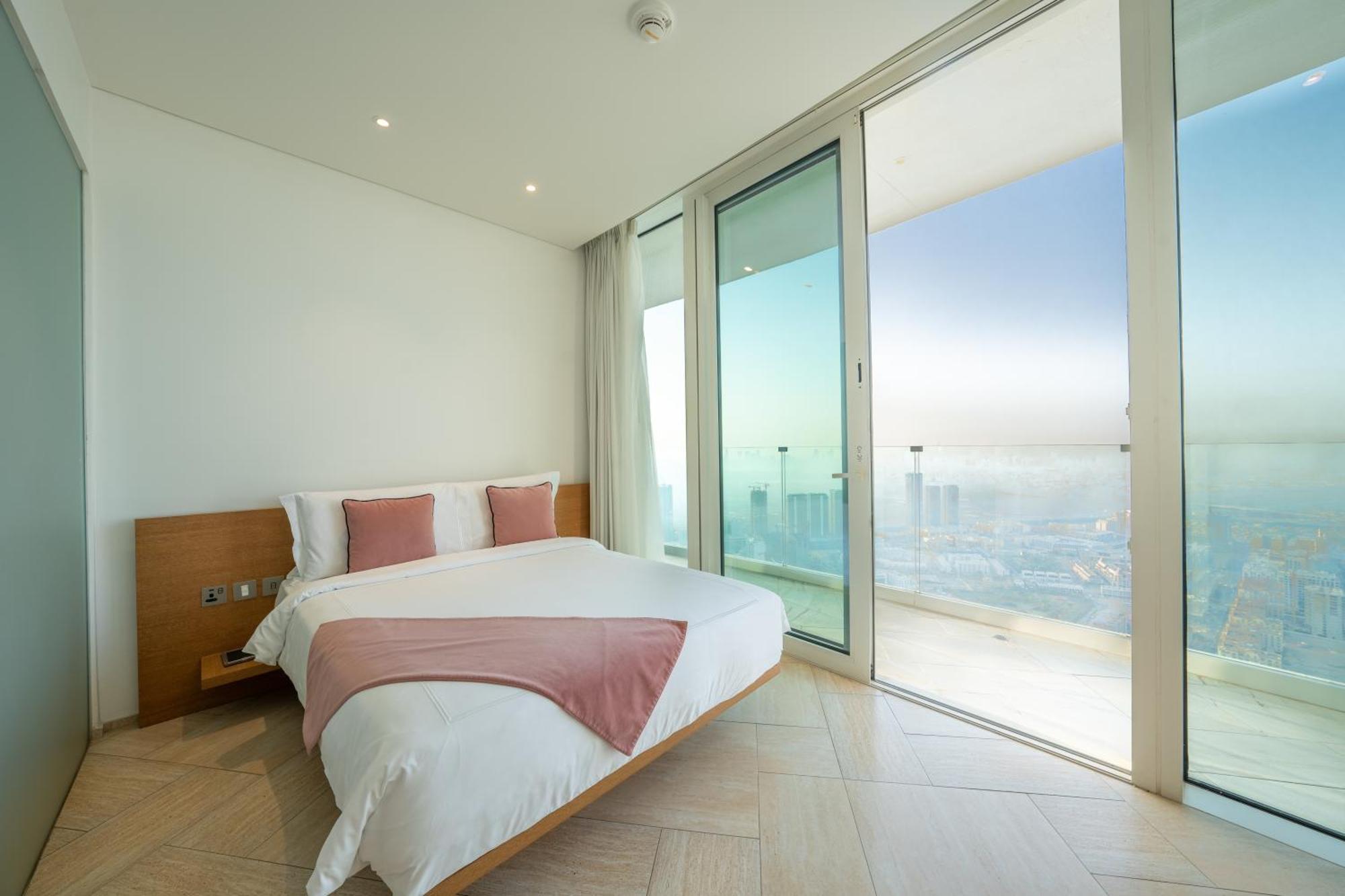 Five Jumeirah Village ดูไบ ภายนอก รูปภาพ The photo shows a modern bedroom with a large bed featuring white bedding and accent pillows in a soft pink color. The room has floor-to-ceiling glass doors that lead out to a balcony, providing a panoramic view of the outside. The walls are painted 