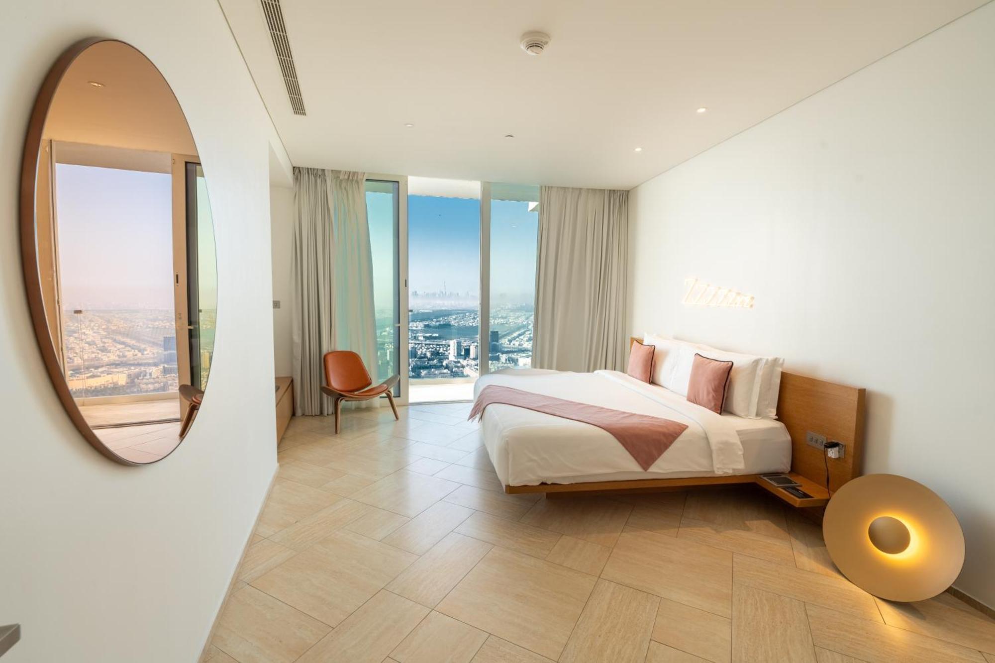 Five Jumeirah Village ดูไบ ภายนอก รูปภาพ The photo shows a modern bedroom with a minimalist design. There is a large bed with white bedding and pink accents, positioned against a wall. To the side, there is a round mirror and a stylish chair. The room features large windows dressed with lig