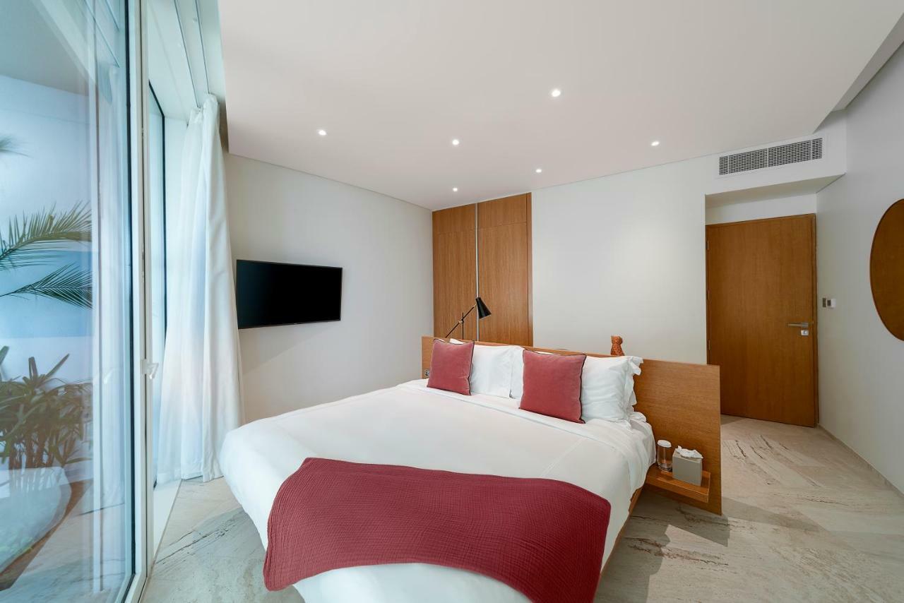 Five Jumeirah Village ดูไบ ภายนอก รูปภาพ The photo shows a modern and stylish bedroom. It features a bed with white bedding and two red decorative pillows. There is a mounted flat-screen TV on the wall opposite the bed. The room has large glass windows that allow natural light to enter, and