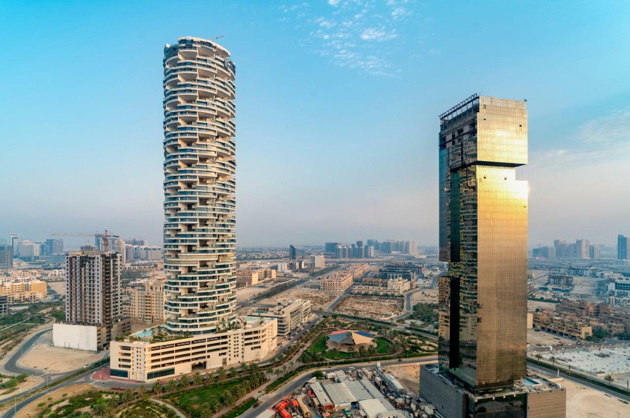 Five Jumeirah Village ดูไบ ภายนอก รูปภาพ The photo shows two distinctive skyscrapers located in an urban area. On the left, there is a tall, cylindrical building with a unique, modern design characterized by its twisting, spiral shape and balconies at various levels. To the right, there is 