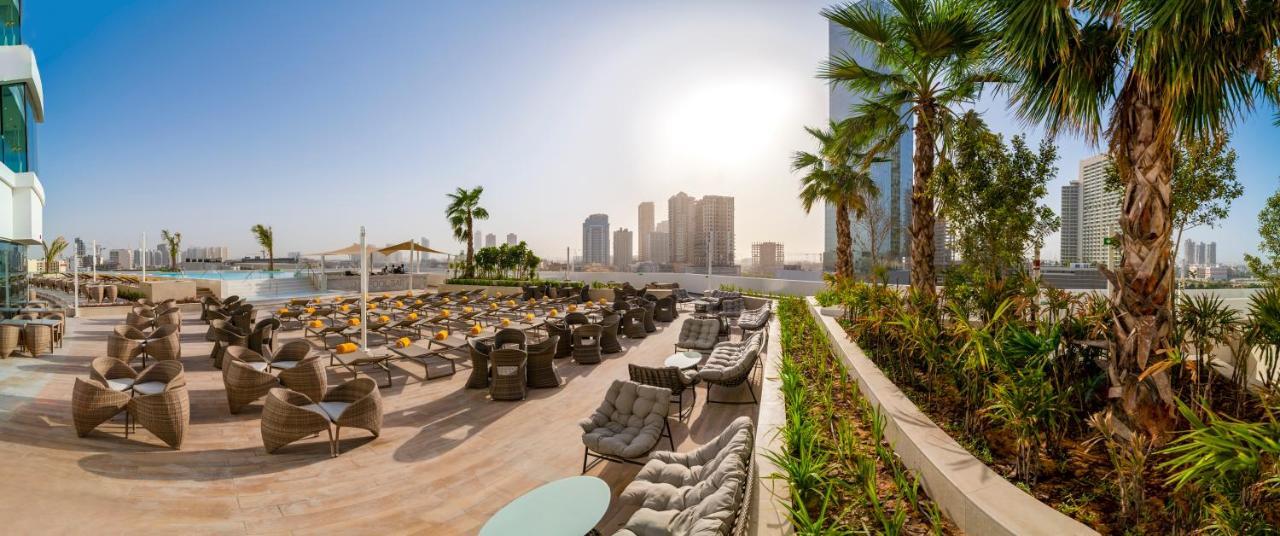 Five Jumeirah Village ดูไบ ภายนอก รูปภาพ The photo showcases a luxurious outdoor seating area, likely part of a resort or hotel. In the foreground, there are well-arranged lounge chairs and seating options, some with cushions, placed on a tiled surface. The area is bordered by lush green pl